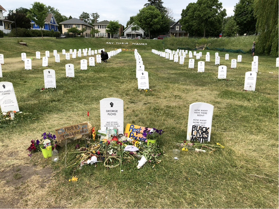 An art installation of with a collection of mock gravestones each bearing the name of a black man killed by police. Say their names is written on the hill in the background.